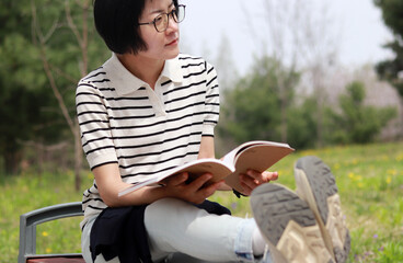A bespectacled woman looking at a book in short-sleeve knitwear on a park bench in warm weather.
