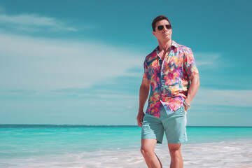 man dressed in a colorful Hawaiian shirt and shorts stands on a sunny beach, exuding relaxed vacation vibes