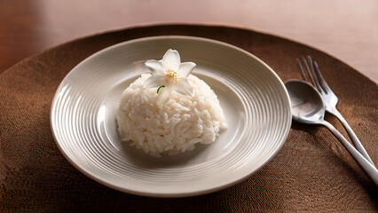 A plate of steamed rice with a small white jasmine flower placed delicately on top. Minimalist composition, soft natural lighting, clean and peaceful atmosphere.