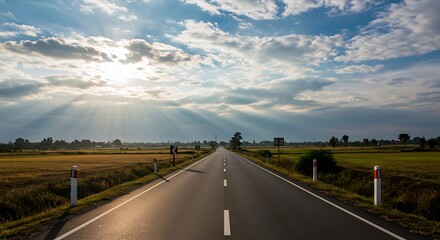 Fototapeta premium A tranquil road meanders through vibrant green fields under a stunning sky filled with clouds. Rays of sunlight illuminate the landscape, creating a serene and picturesque atmosphere at dusk