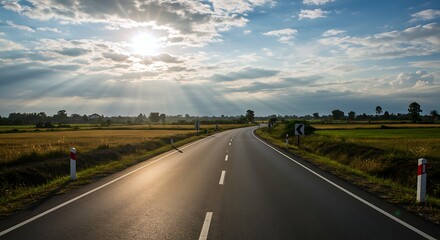 Fototapeta premium A winding road stretches through lush countryside, illuminated by golden sunlight. Fields of crops sway gently in the breeze beneath a dramatically lit sky at dusk