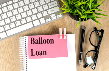 Notebook with pink note reading balloon loan on wooden desk with keyboard, pen, eyeglasses and...