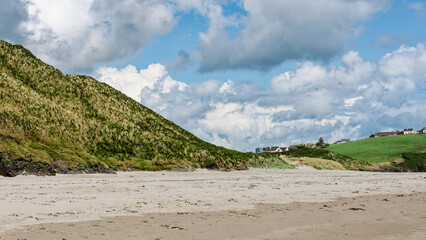 Idyllic coastal setting. A scenic beach backed by rolling green hills and charming houses. A calm atmosphere with a few people enjoying the day.