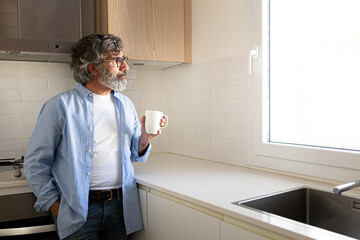 Pensive mature man relaxing at home having morning coffee looking out the kitchen window. Copy space.