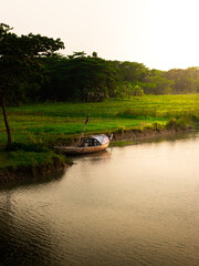 Fototapeta premium Traditional wooden boat anchored by the riverbank at sunset, surrounded by lush green fields and dense trees, reflecting the serene beauty of rural life.