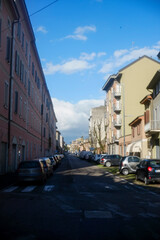 Fototapeta premium Cars are parked along a narrow street lined with colorful buildings, under a vibrant blue sky dotted with white clouds, creating a picturesque urban scene