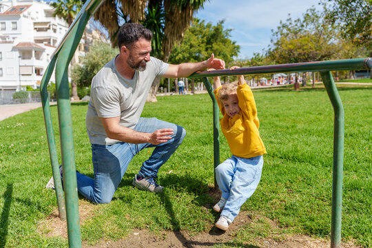 Father kneeling on lush grass, helping smiling son hang from monkey bars in a vibrant, sunny park filled with joy and laughter - Powered by Adobe