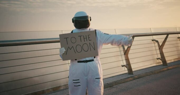 Female Astronaut hitchhiker in Space Suit with cardboard poster with words TO THE MOON. Hispanic woman cosmonaut exploring deep cosmos. Cyber punk aesthetics