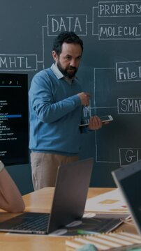 Medium vertical shot of middle-aged Caucasian male coding teacher standing in front of blackboard with algorithm scheme, interacting with offscreen students while conducting lesson