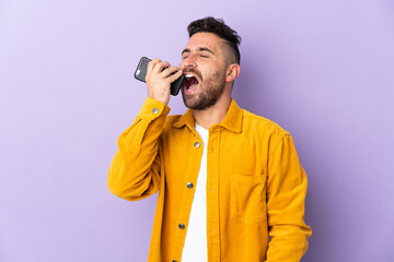 Naklejka premium Caucasian man isolated on purple background using mobile phone and singing