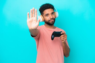 Young brazilian man playing with a video game controller isolated on blue background making stop gesture © luismolinero