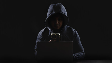 Suspicious man wear hoodie using computer laptop and smartphone to hack in to system in the dark...