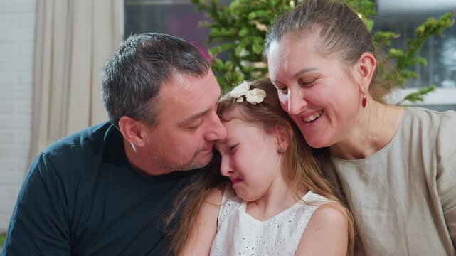 Close up view of loving dad trying to kiss young crying daughter on cheek while smiling mom looks on warmly during touching family moment near christmas tree inside cozy festive decorated home