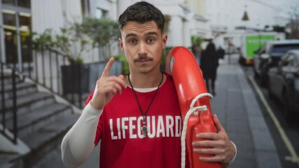 Young hispanic male lifeguard in red shirt outdoors holding buoy on crowded city street.