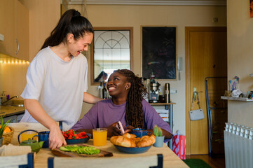 Happy interracial lesbian couple sharing a joyful breakfast in a cozy kitchen, surrounded by fresh fruits and pastries, fostering love and connection