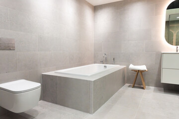 Modern built-in ceramic bathtub with stainless steel and chrome faucets in a contemporary bathroom with gray tile walls and floor for elegant home interior concept