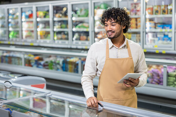 A grocery store employee smiles, using a tablet while checking the refrigerated goods section in...