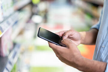 A person holding a smartphone in a supermarket aisle, possibly shopping or comparing prices. The soft focus highlights the technology and the setting.