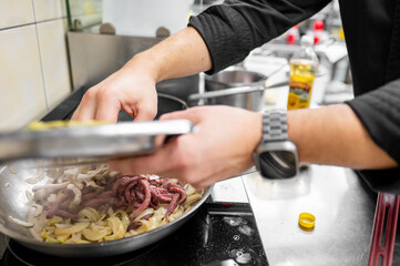 A chef stirs a skillet filled with sautéed onions and strips of meat. The engaging kitchen environment is alive with steam, highlighting the cooking process in a professional setting.