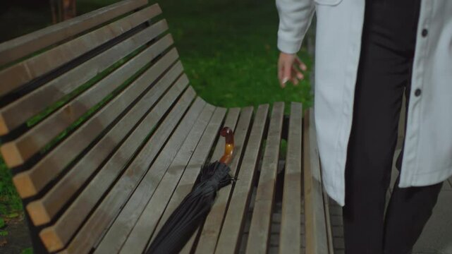 Lady dropping umbrella on wooden bench holding book, preparing to sit down and read during calm night in outdoor park setting, surrounded by greenery and illuminated by soft streetlight glow