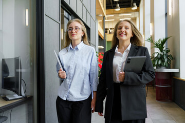 Two Professional Women Confidently Walk Through a Modern Office Space With Laptops and Tablets in Hand