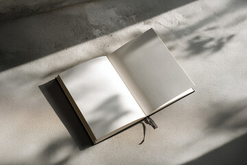 Flat lay of a blank sketchbook placed on a textured concrete floor with dramatic natural light and shadow patterns. A moody and artistic setting, great for mockups related to drawing, note-taking, or 