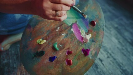 Experienced painter carefully selecting green oil paint, loading brush from wooden palette filled with vibrant colors while preparing to create artistic composition