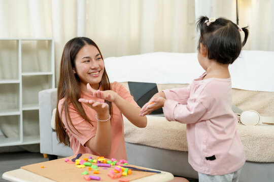 Asian single mother smiling while clapping hands with toddler daughter during clay playtime on table expressing encouragement and connection in creative learning at home in happy bonding moment