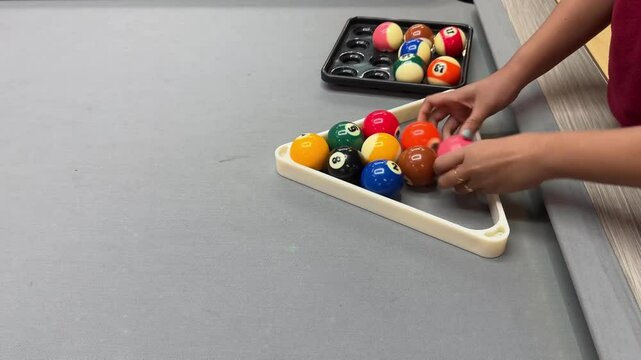 Player arranging colorful billiard balls into a triangle rack on a gray pool table, preparing for a game. Focus on hands, balls, and setup process in a casual indoor setting.