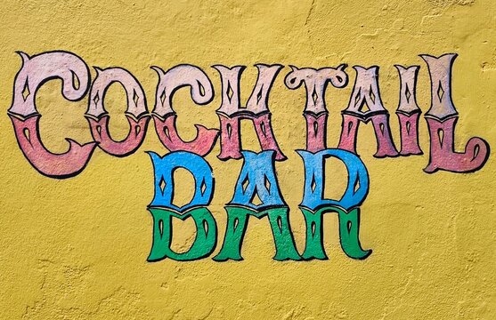 Photo of a vibrant "Cocktail Bar" sign painted on a bright yellow wall, featuring Wild West-style saloon lettering in bold pink, blue, and green hues that pop with playful, retro charm.