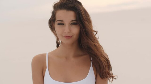 A beautiful young woman in a white outfit poses shyly while looking at the camera. She smiles sweetly and plays with her hair, creating a soft, natural, and charming moment. 