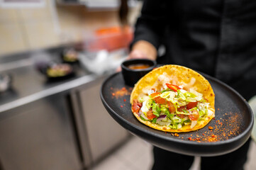 A chef presents a vibrant taco filled with fresh lettuce, diced tomatoes, and a side of dipping sauce. The kitchen backdrop showcases a lively atmosphere, perfect for culinary creativity.