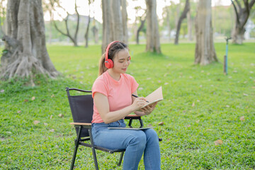 Asian woman sitting listening to music, reading a book, outdoors in nature, evening.