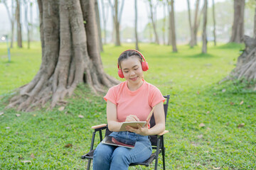 Asian woman sitting listening to music, reading a book, outdoors in nature, evening.
