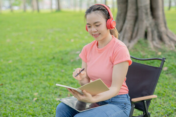 Asian woman sitting listening to music, reading a book, outdoors in nature, evening.