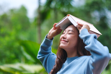 Portrait image of a woman with closed eyes holding and putting a book on her head in the garden