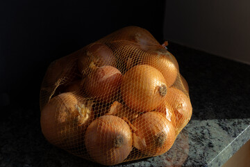 Yellow onions in mesh produce bag on green granite counter top in sunlight.