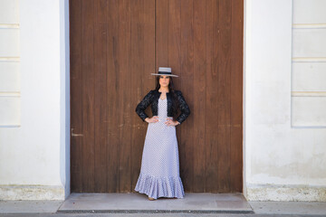 Young woman, beautiful, brunette, flamenco dancer, wearing beautiful dress with jacket and hat, posing with wooden door in background. Flamenco concept, dancer, typical, Spanish, Seville.