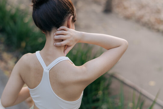 Asian woman experiencing neck pain after exercise, wearing white sports bra, standing outdoors in park with greenery in background, expressing discomfort