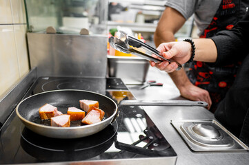 A chef uses tongs to flip chunks of meat sizzling in a frying pan on a stovetop. The busy kitchen environment is lively, showcasing the art of cooking in progress.