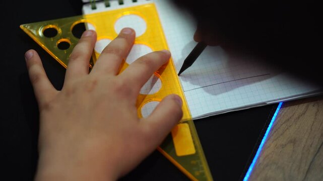 boy draws simple drawing of house with pencil and ruler in squared notebook. hand with pencil and triangle close-up