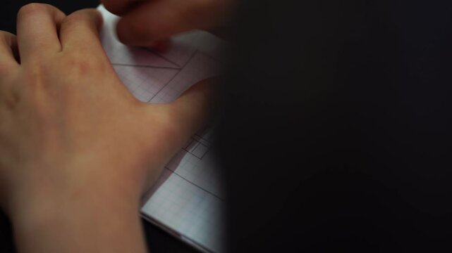boy draws simple drawing of house with pencil and ruler in squared notebook. hand with pencil and triangle close-up