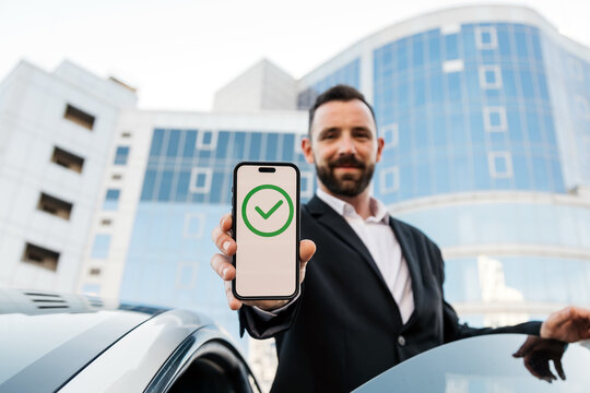 A businessman holds a phone next to his modern car with a sign of approved transfer or payment on the screen. The concept of confirmed, successful, completed or approved status.
