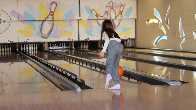 A young player focuses on the bowling lane, holding an orange ball and preparing for her throw in a vibrant bowling alley filled with colorful decor
