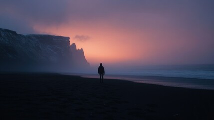 Fototapeta premium A lone figure silhouetted against a dramatic sunset over a misty, snow-dusted Icelandic beach.