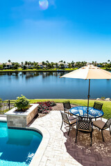 Fototapeta premium glass-top patio table with metal chairs sits under a beige umbrella overlooking a tranquil lake and pool.