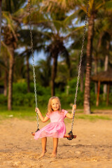 adorable caucasian little girl swinging swings on sandy beach at hot summer day. Child having fun during tropical vacation on sea