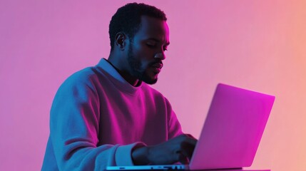 A young man sits in front of a laptop, illuminated by vibrant pink and blue neon lights.
