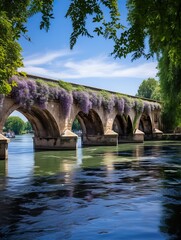 Fototapeta premium Stone bridge adorned with wisteria over a river under a blue sky with green foliage framing the scene