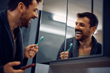 Cheerful man brushing teeth while looking himself in mirror.